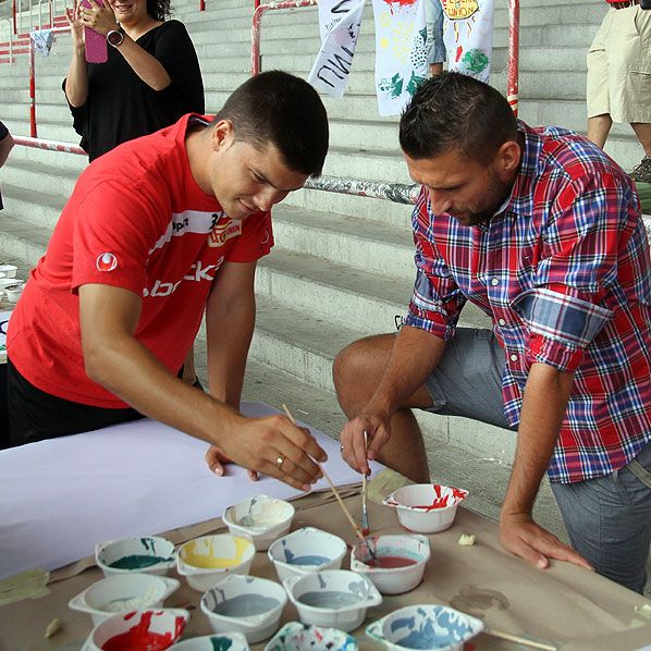 Zwei Männer stehen an einem Tisch und malen mit Pinseln in bunten Farben, während im Hintergrund eine Person lächelt.