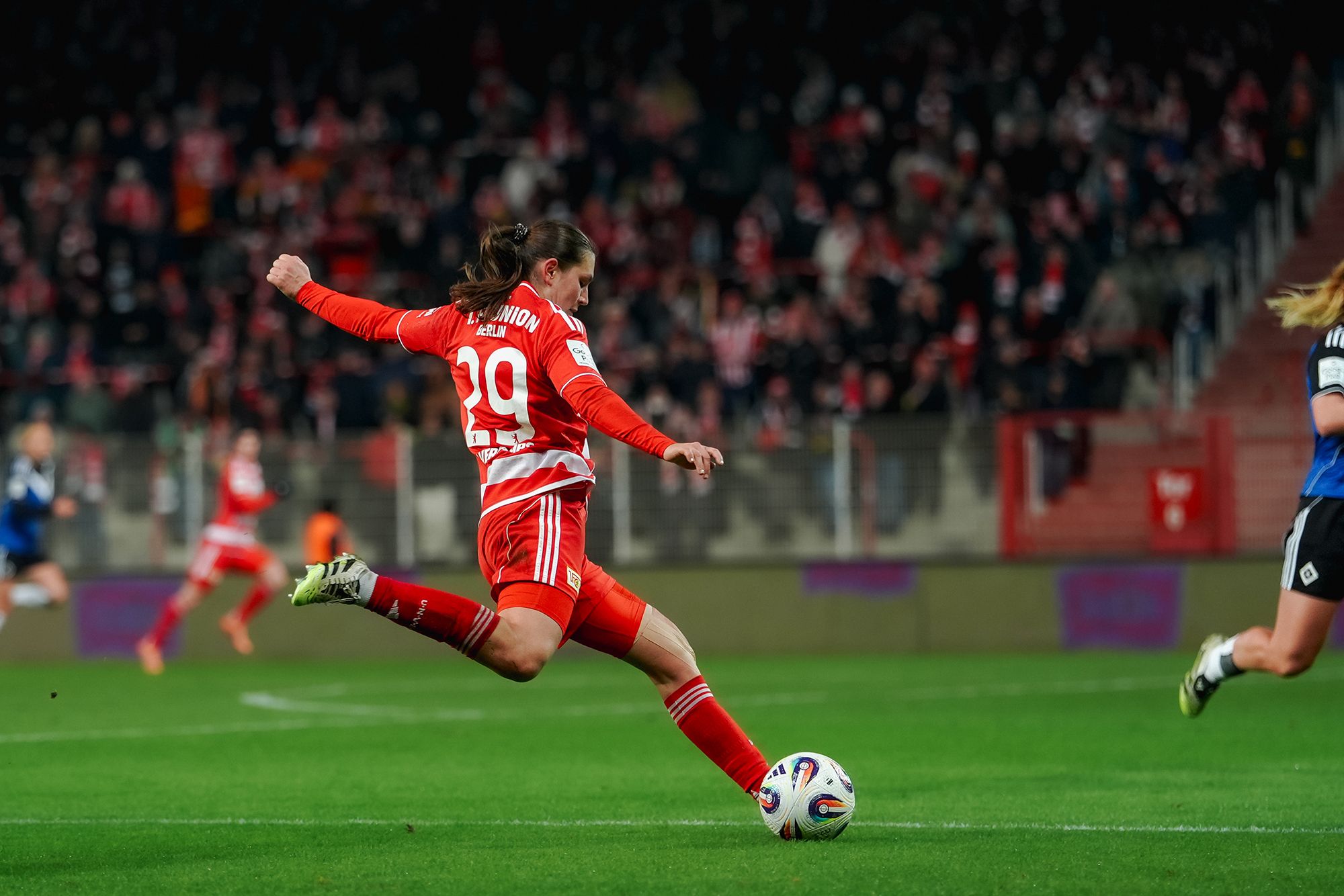 Spielerin in roter Sportkleidung schießt den Ball während eines Fußballspiels vor einer jubelnden Menge.