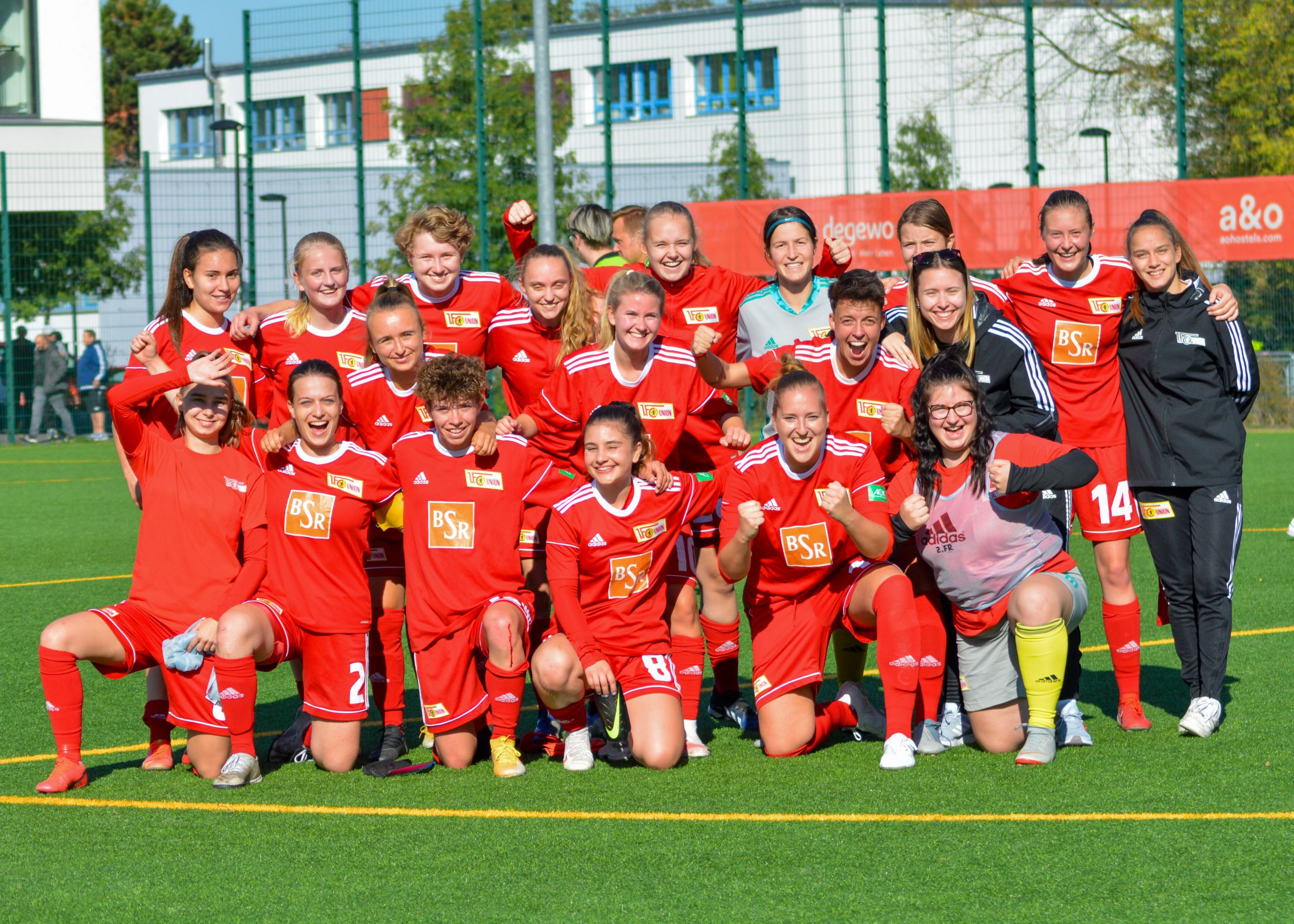 Gruppenselfie von einer Frauenfußballmannschaft in roten Trikots auf einem Sportplatz, alle lächeln und posieren zusammen.