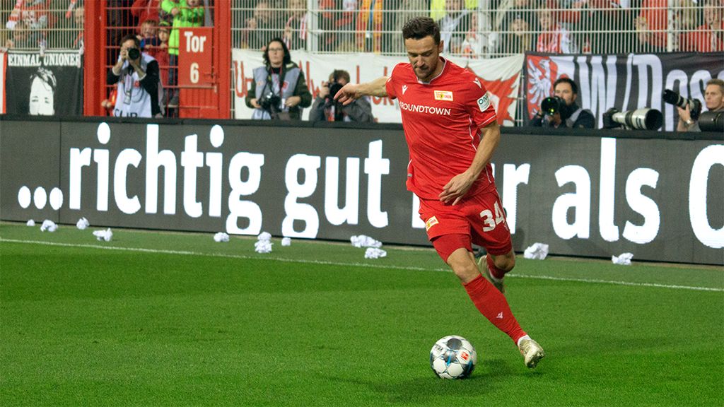 Ein Fußballspieler trägt ein rotes Trikot. Er dribbelt den Ball. Im Hintergrund sind Zuschauer.
