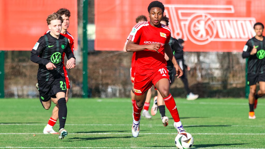 Ein Fußballspieler trägt ein rot-weißes Trikot. Er läuft schnell über das Spielfeld. Im Hintergrund sind Spieler in schwarz-grünen Trikots.
