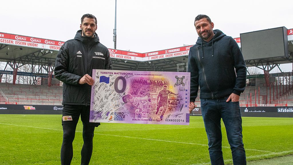 Zwei Männer stehen auf einem Fußballfeld und halten einen großen Geldschein mit sportlichem Motiv in der Hand.