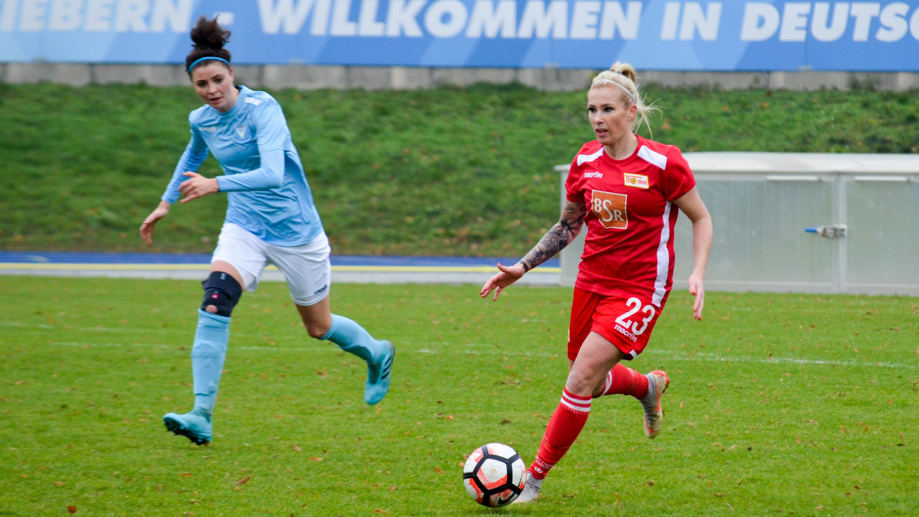 Zwei Fußballspielerinnen spielen. Eine trägt rot, die andere blau. Sie laufen dem Ball nach.