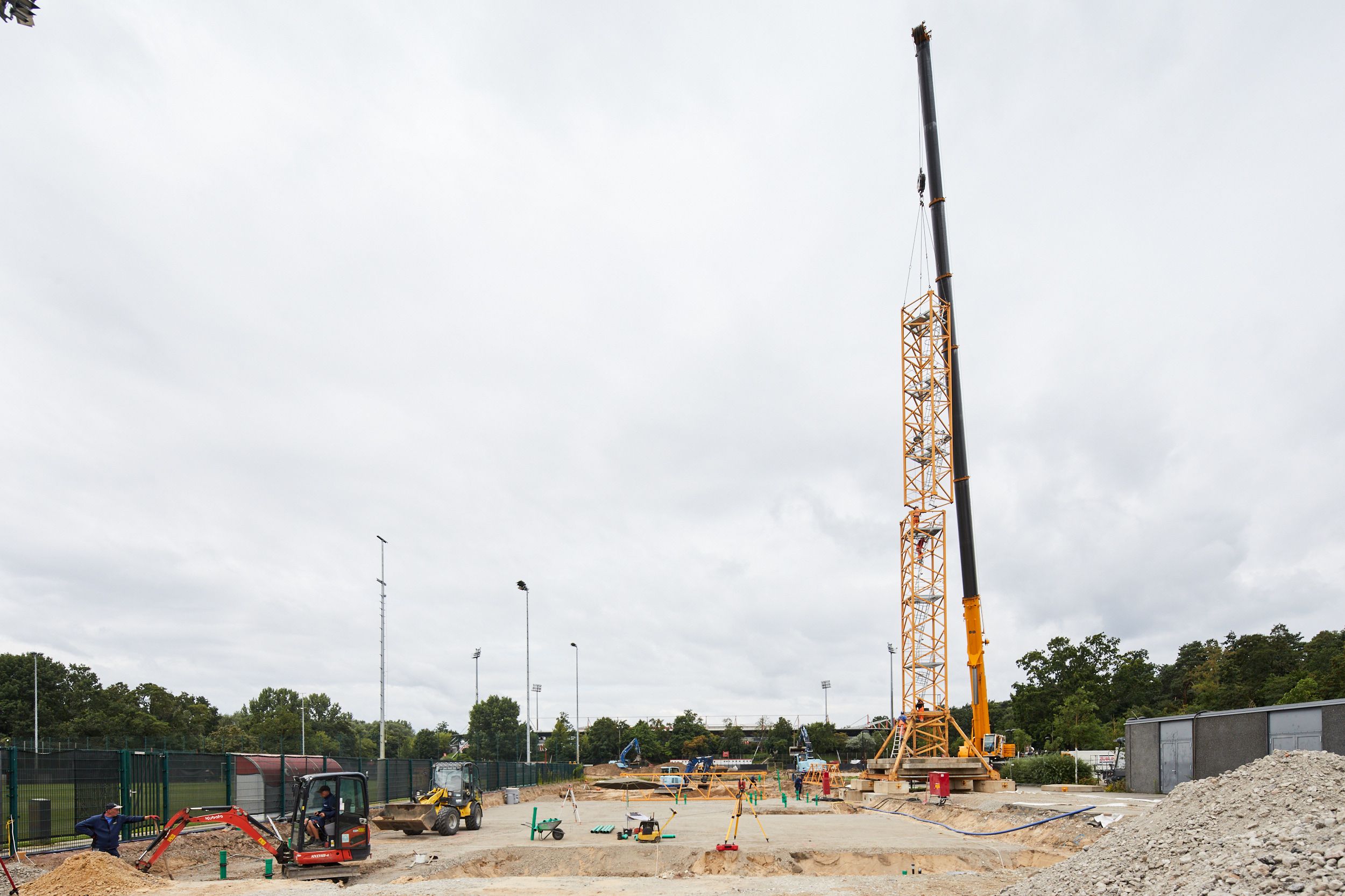 Construction site with crane, earth-moving machines, and surrounding construction materials under a cloudy sky.