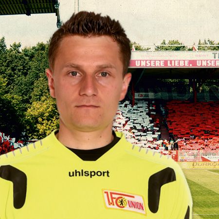 Ein Fußballtorwart in gelber Spielkleidung vor einem Stadion mit Fans und Teamfarben im Hintergrund.
