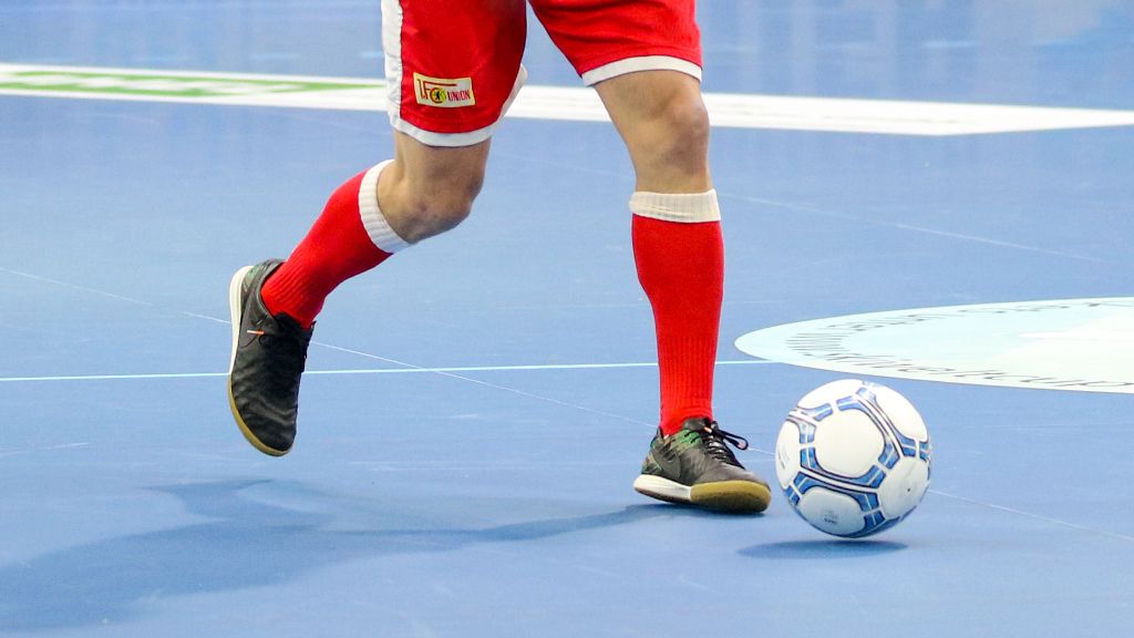 Ein Spieler in roten Shorts und Socken tritt einen Fußball auf einem blauen Spielfeld.