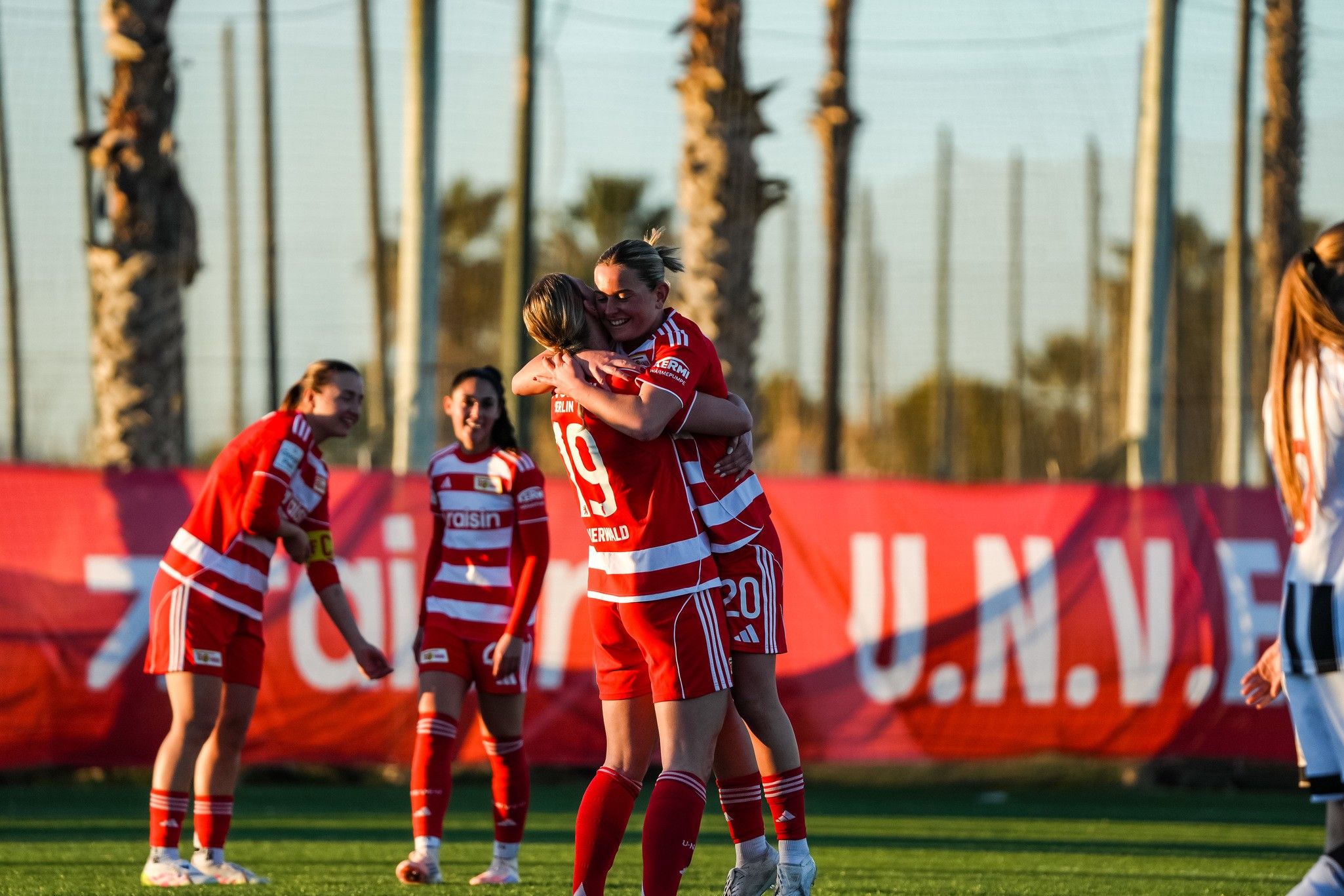 Zwei Fußballspielerinnen in roten Trikots umarmen sich jubelnd auf dem Spielfeld, während andere Teamkolleginnen im Hintergrund stehen.