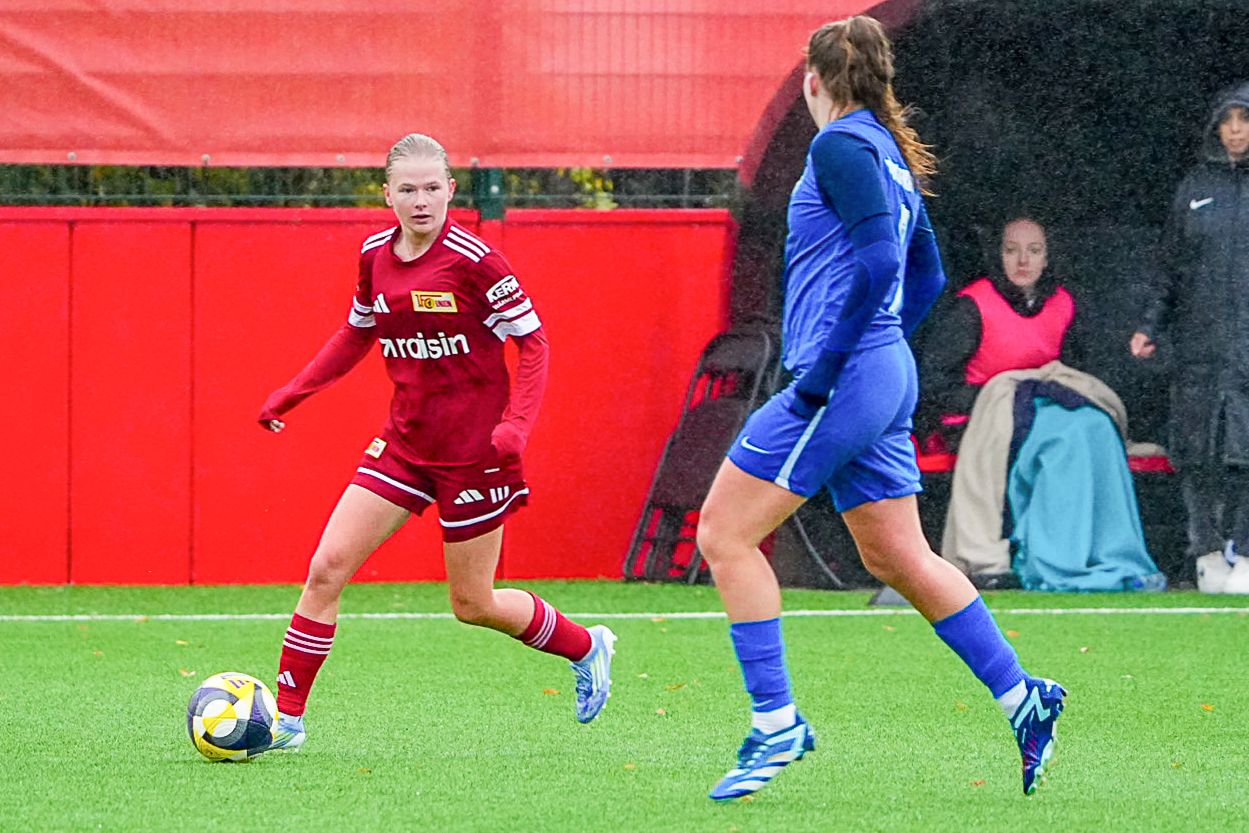 Eine Fußballspielerin in rotem Trikot dribbelt den Ball auf einem Platz, während eine andere Spielerin in blauem Trikot defensiv steht.