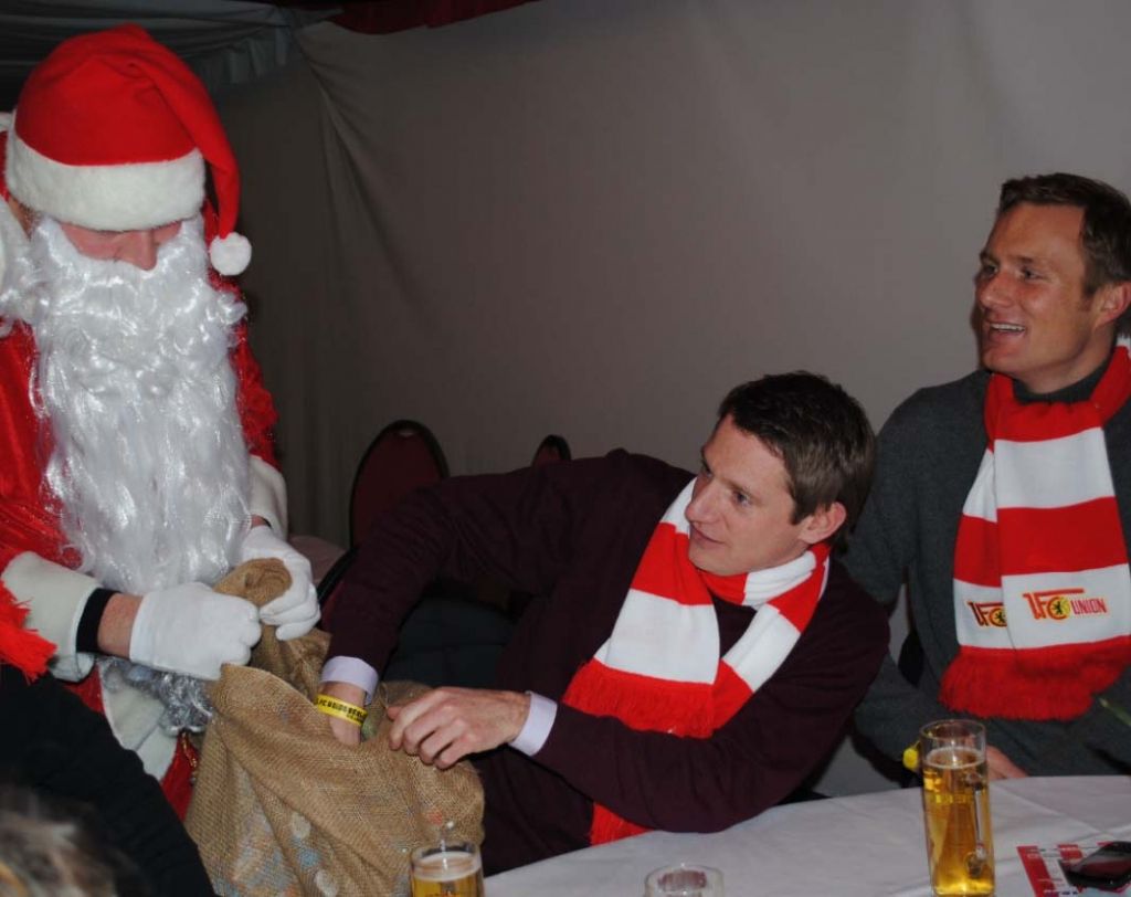 Ein Weihnachtsmann überreicht einem Mann in einem roten Schal eine Geschenktüte, während ein weiterer Mann daneben sitzt.