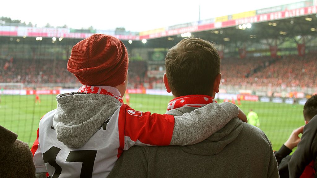 Two football fans are in a stadium watching the game, one wearing a cap and the other has his arm around him.