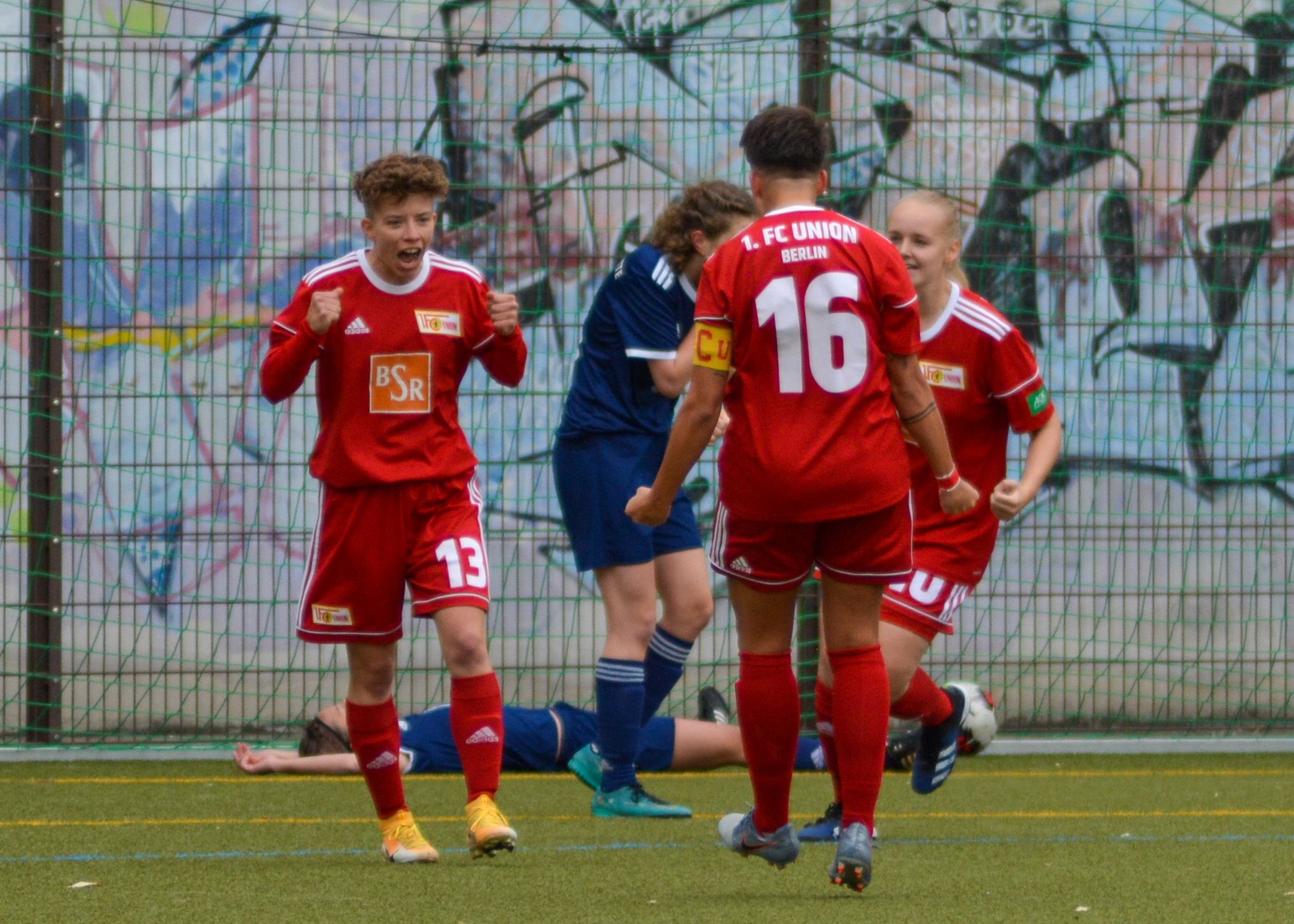 Junge Fußballspieler in roten Trikots jubeln auf dem Spielfeld vor einer Graffiti-Wand, während ein Gegner am Boden liegt.