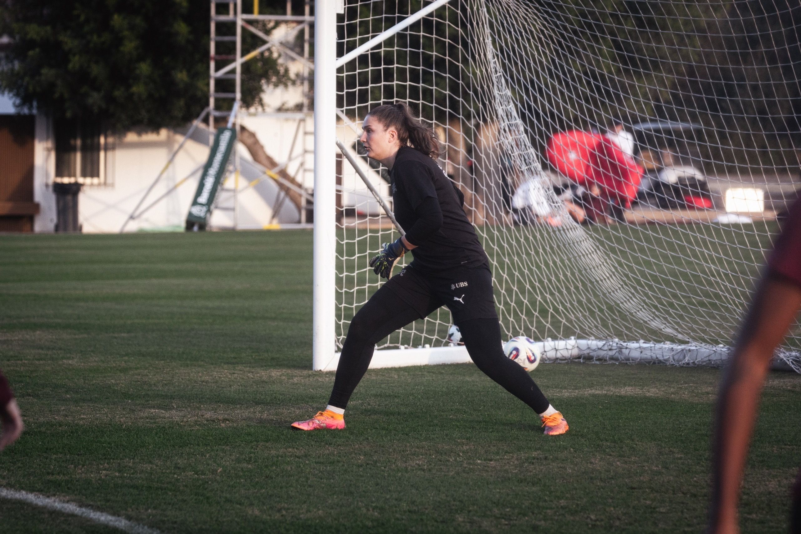 Eine Torhüterin steht bereit im Tor, während das Spiel auf einem Fußballfeld im Hintergrund stattfindet.