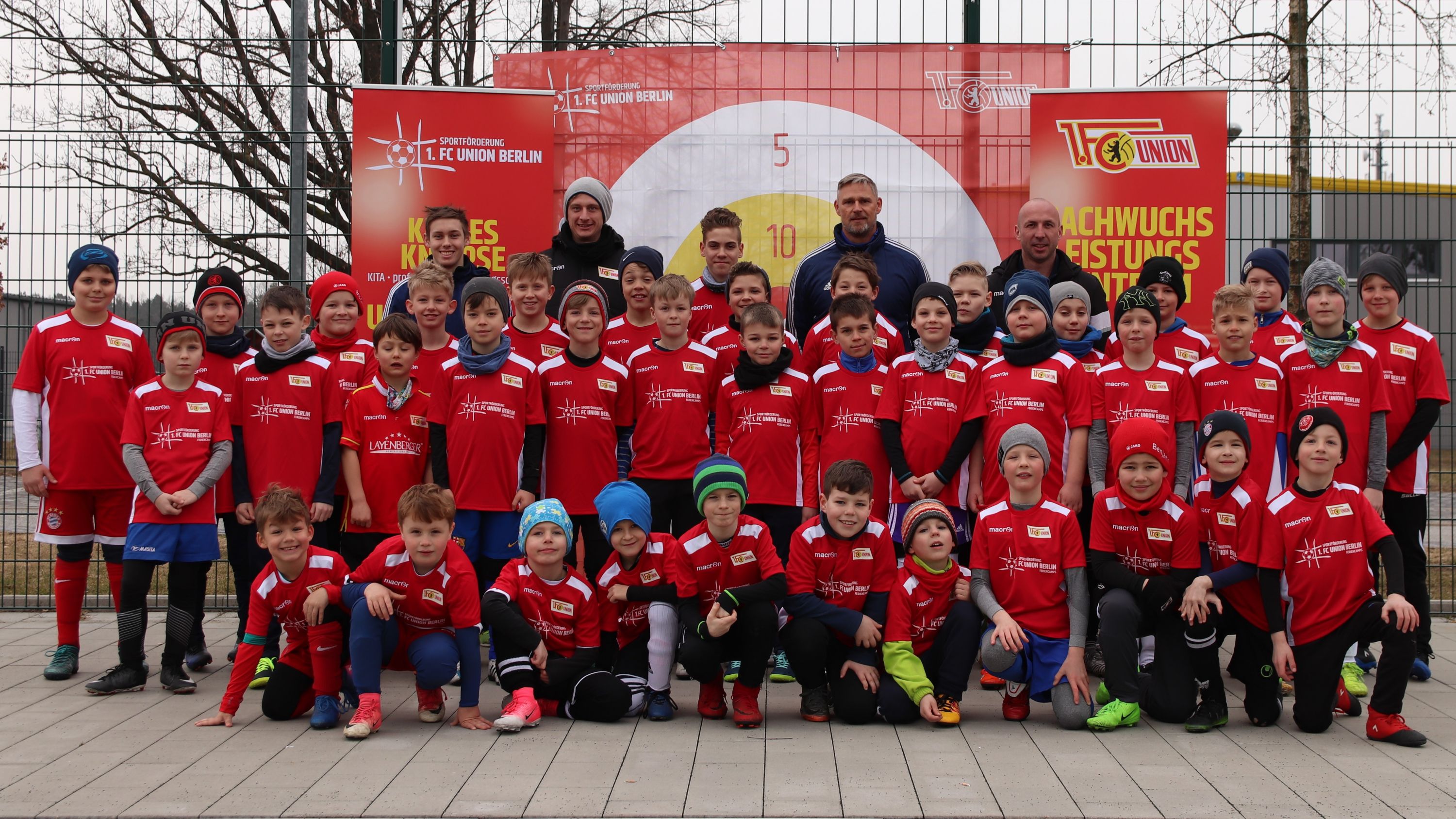 Gruppenfoto von 50 Jungen in roten Trikots, die auf einem Sportplatz stehen, mit einem Banner im Hintergrund.