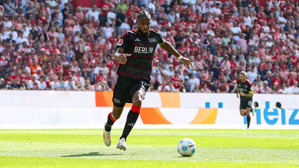 A soccer player in a black uniform with "BERLIN" written on it dribbles the ball on a green field in front of a cheering crowd.