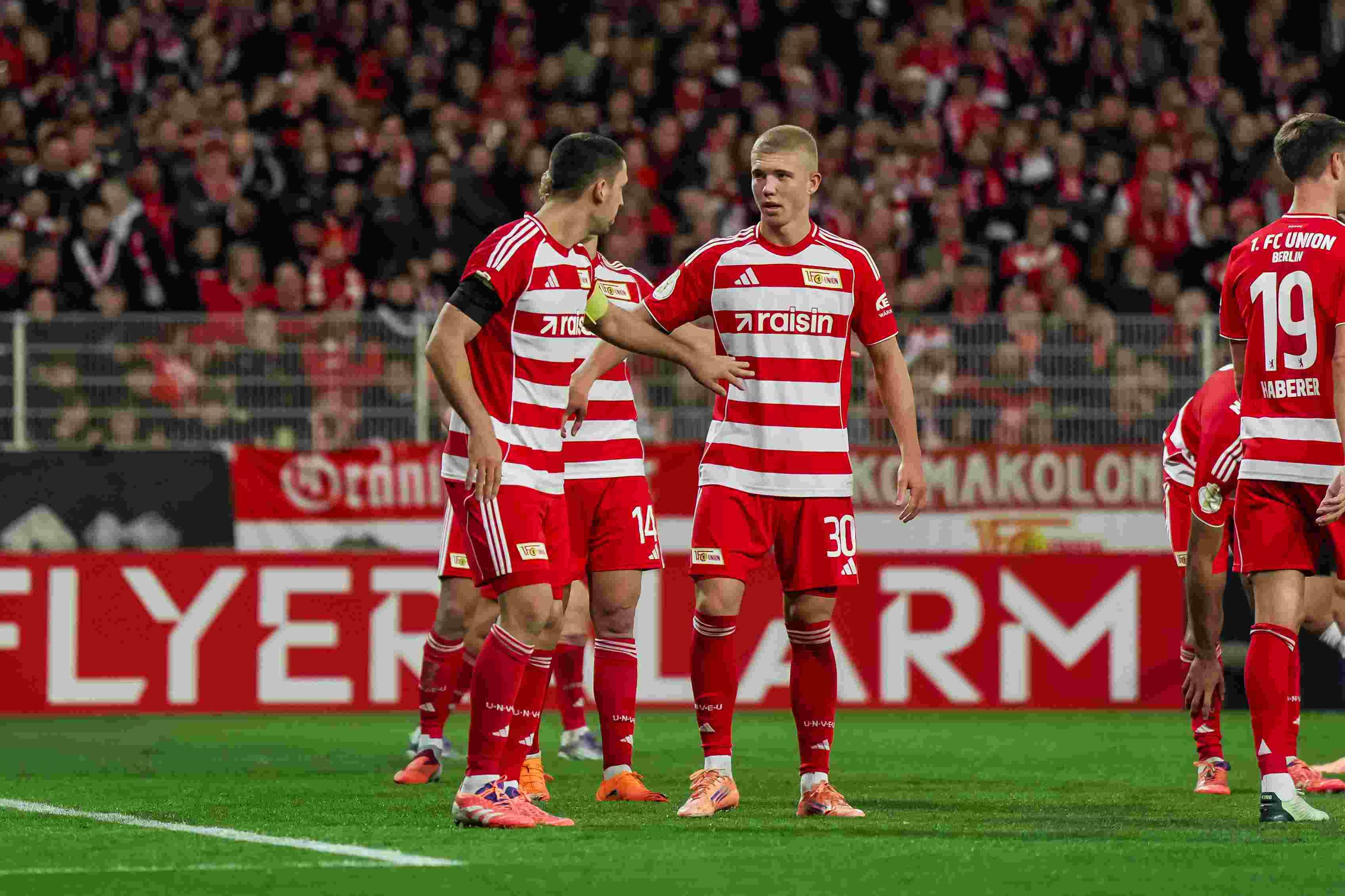 Zwei Fußballspieler in roten und weißen Trikots stehen auf dem Feld und sprechen miteinander, im Hintergrund Fans im Stadion.