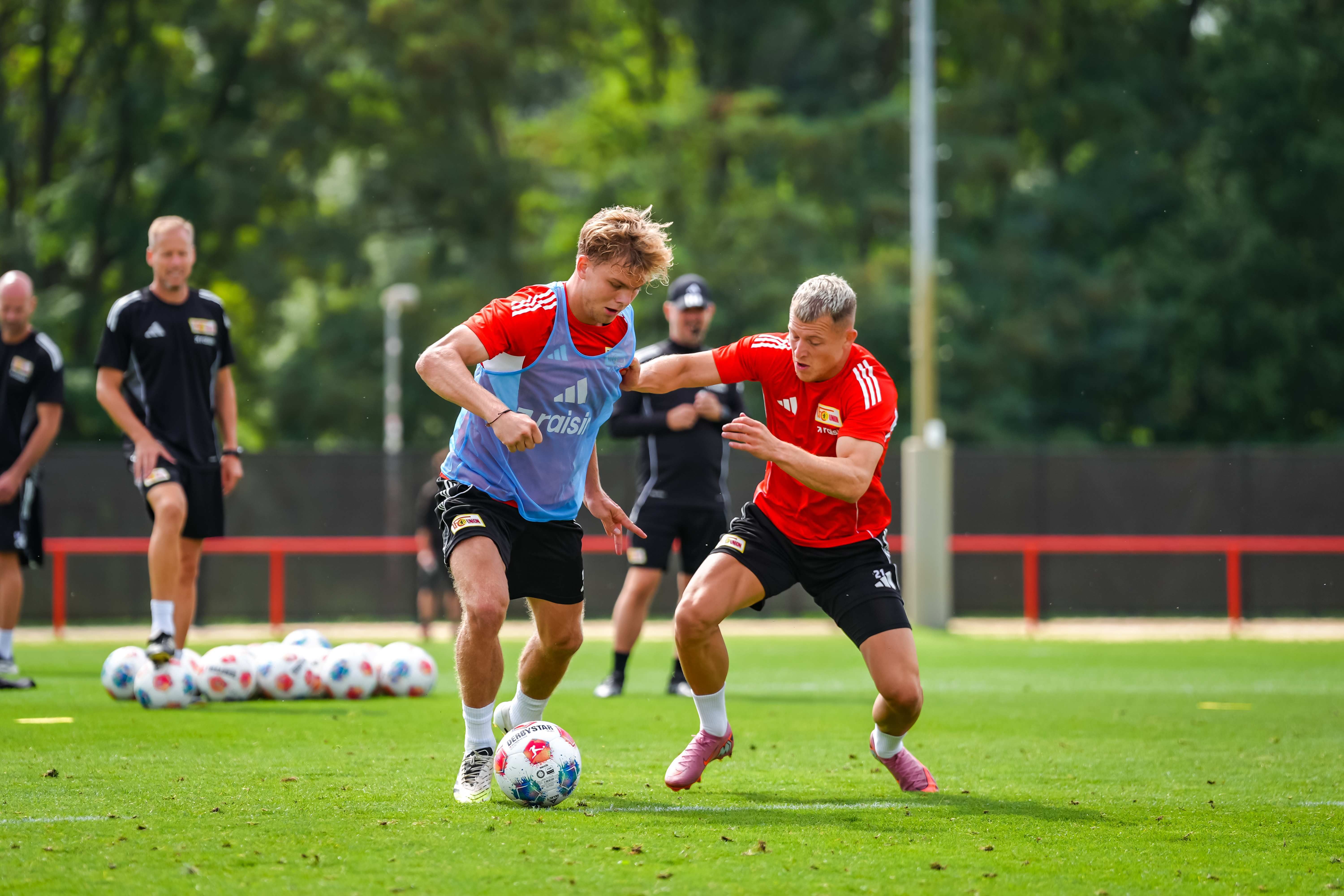 Two soccer players are in training, one is dribbling the ball while the other tries to stop it. Background with other players.