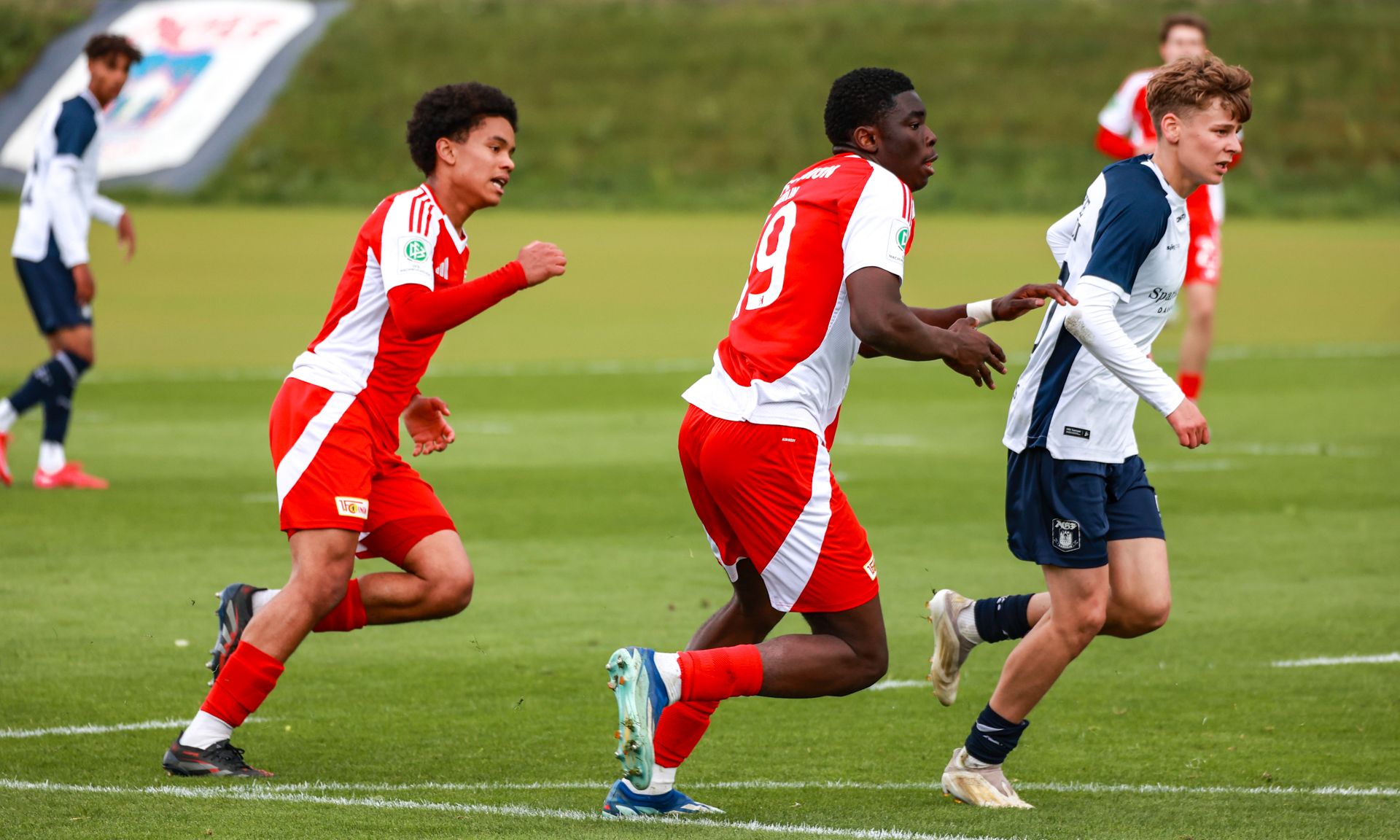 Drei Fußballspieler tragen rote und weiße Trikots. Ein Spieler trägt ein dunkelblaues Trikot. Sie laufen auf dem Rasen.