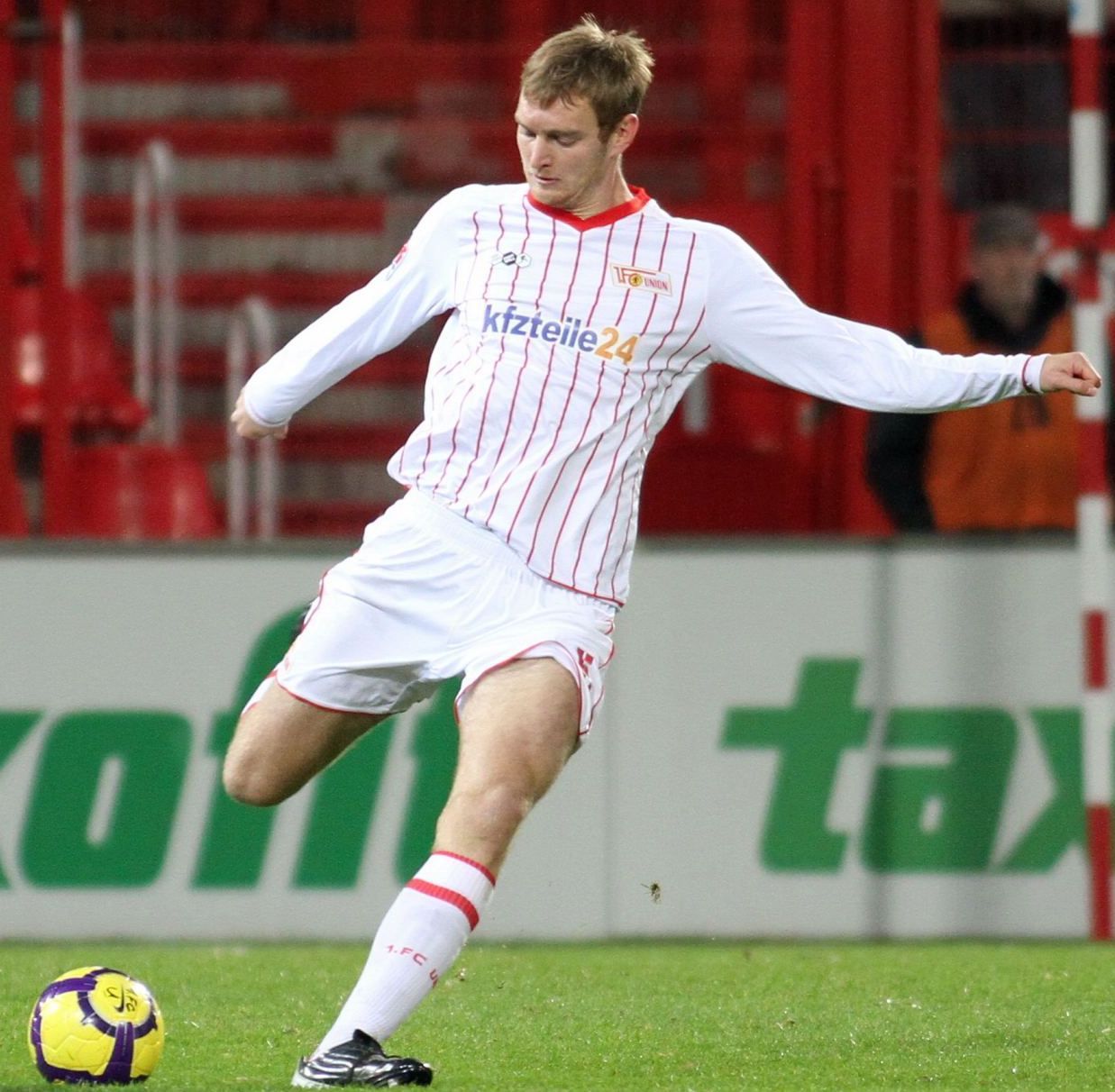 Ein Fußballspieler in einem weißen Trikot mit roten Streifen, der den Ball auf einem Stadionrasen spielt.