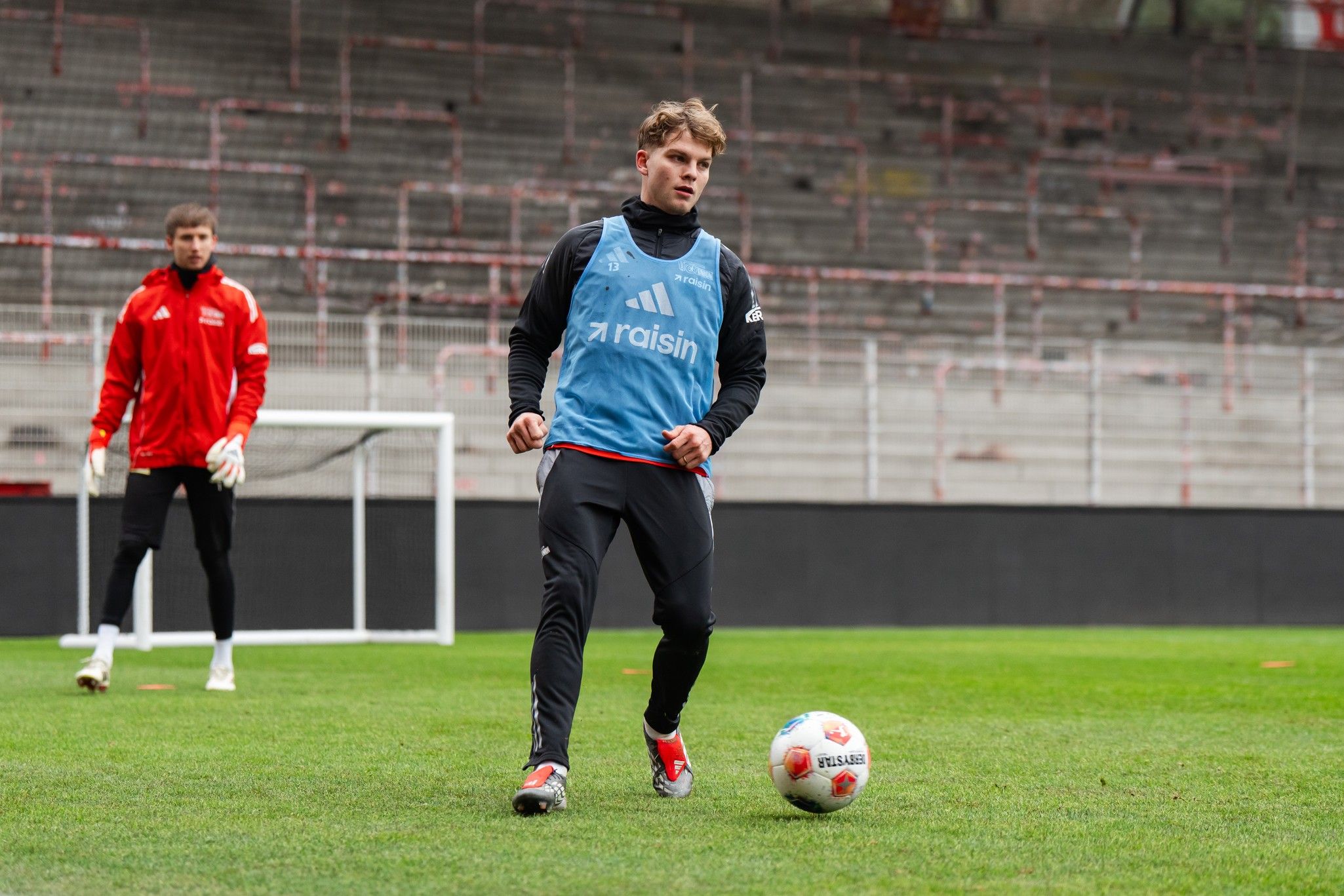 Ein Fußballspieler trainiert auf einem Rasenplatz, während ein Torwart im Hintergrund steht.