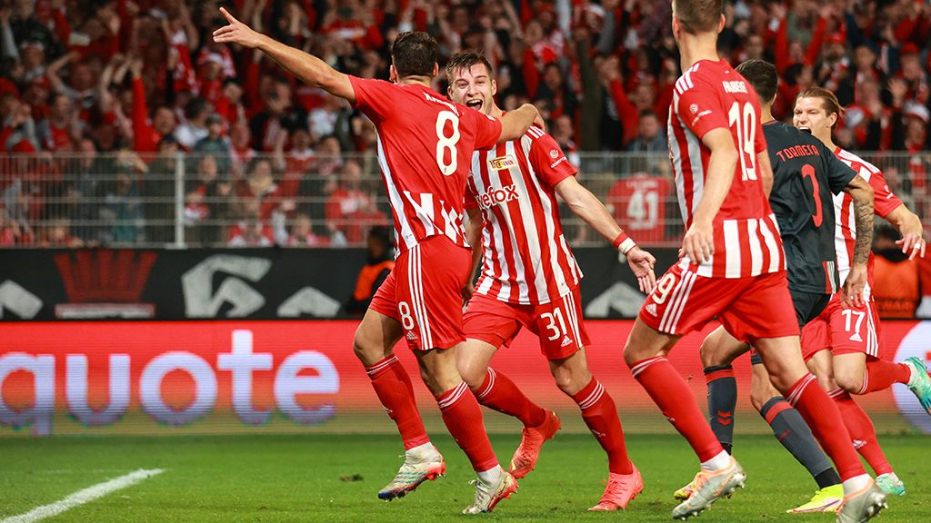 Two soccer players in red jerseys celebrate a goal among cheering fans in the stadium.