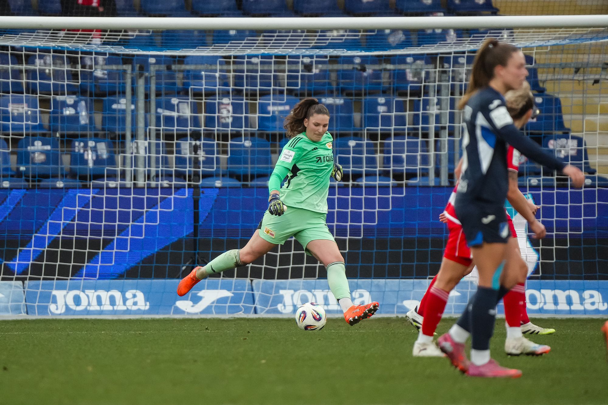 Torhüterin in grüner Ausrüstung spielt den Ball im Fußballspiel, während Spielerinnen in blauen und roten Trikots im Hintergrund agieren.