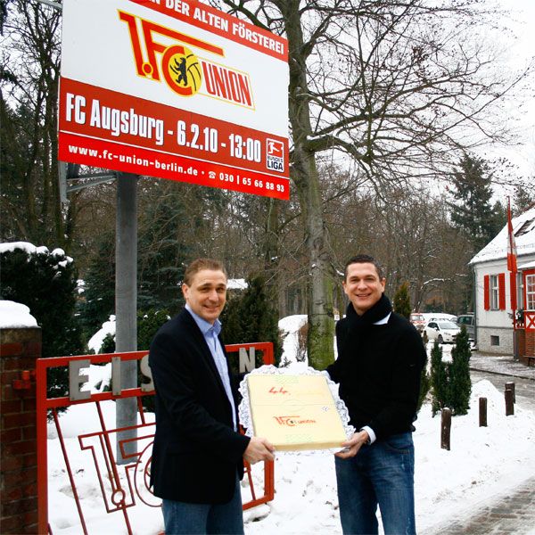 Zwei Männer stehen im Schnee vor einem Schild des FC Union Berlin und halten eine große Torte.
