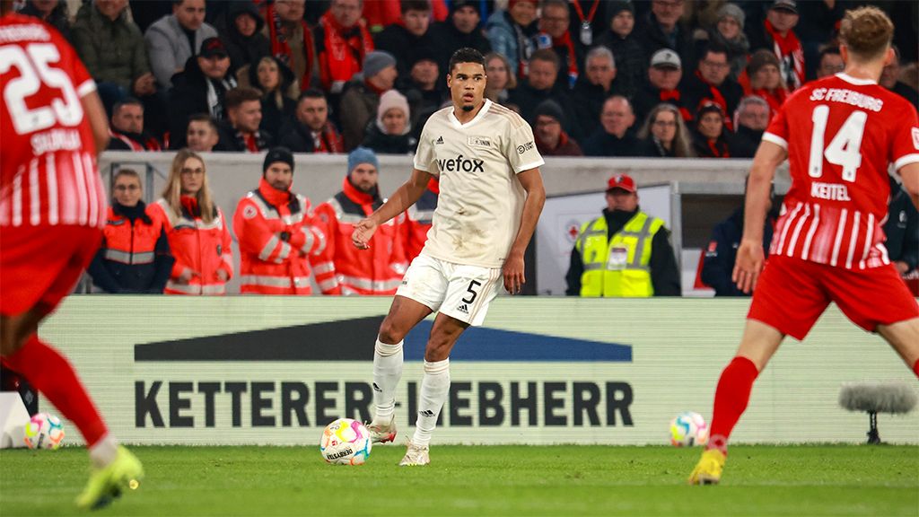 Ein Fußballspieler in einem hellen Trikot steht auf dem Spielfeld, während zwei Gegner in roten Trikots sich ihm nähern.
