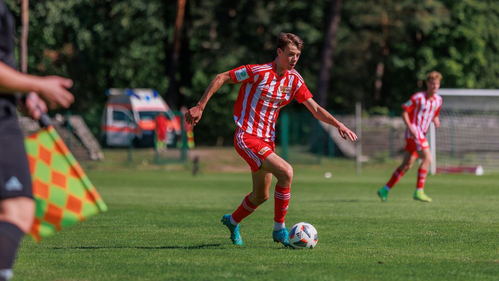 Spieler in rot-gestreifter Fußballuniform dribbelt den Ball auf einem grünen Rasenfeld. Im Hintergrund sind Bäume zu sehen.