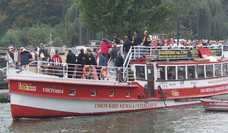 Ein Ausflugsschiff mit vielen Passagieren fährt auf einem Gewässer, umgeben von Bäumen und Grün.