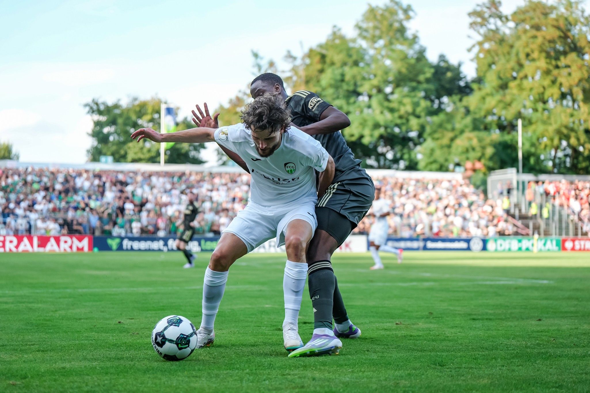 Ein Fußballspieler im weißen Trikot wird von einem gegnerischen Spieler im grauen Trikot bedrängt, während er einen Ball verfolgt.