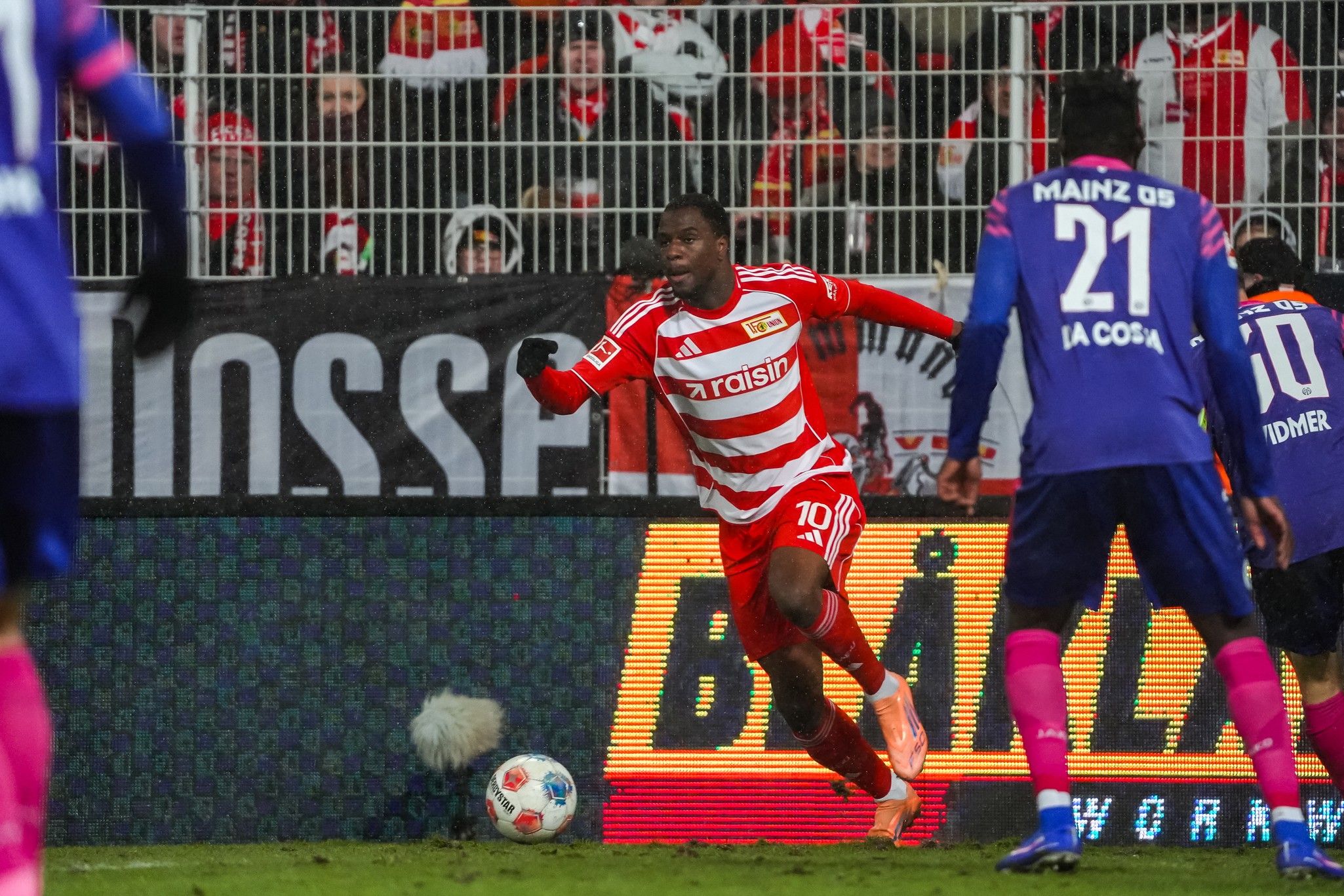 Spielszene im Fußball: Ein Spieler in rotem Trikot sprintet mit dem Ball, während gegnerische Spieler in blauen Trikots ihm folgen.
