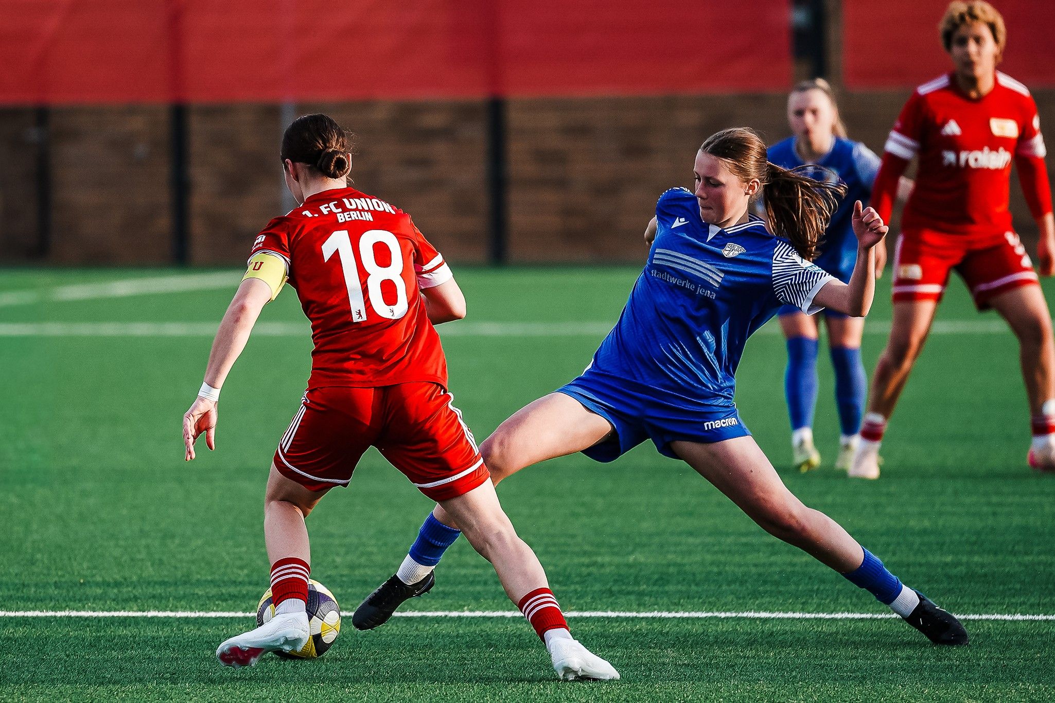 Zwei Frauenfußballspielerin im Wettkampf um den Ball auf einem Kunstrasenplatz. Der Spielerin in Rot ist die Nummer 18 zugeordnet.