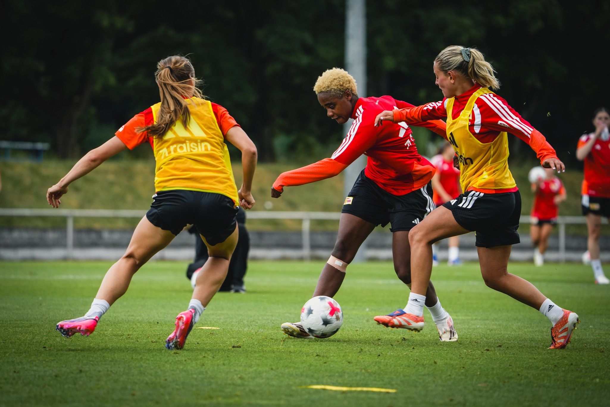 Drei Fußballspielerinnen im Training, die um den Ball kämpfen, auf einem Fußballplatz mit Rasen und Bäumen im Hintergrund.