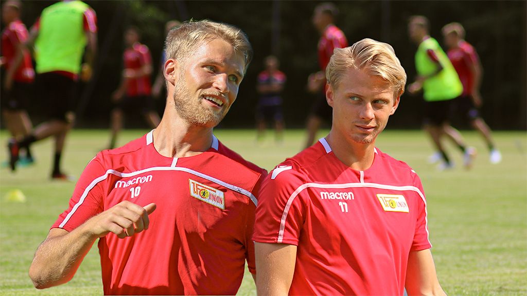 Two football players in red jerseys are standing on a training field and talking. In the background, other players can be seen.
