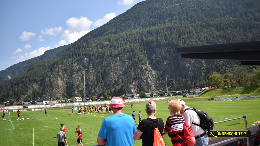 Eine Gruppe von Menschen beobachtet ein Training auf einem Fußballplatz, umgeben von Bergen und klarem Himmel.