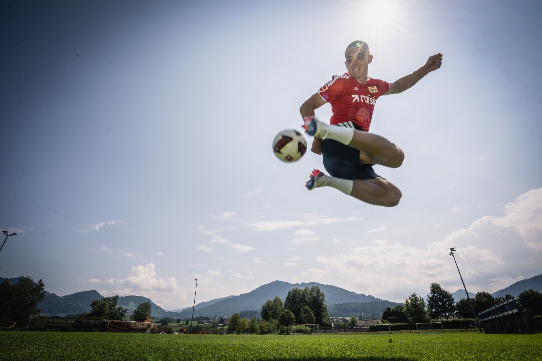 Ein junger Fußballspieler springt im Training hoch in die Luft und führt einen Schuss aus, mit einer sonnigen Landschaft im Hintergrund.