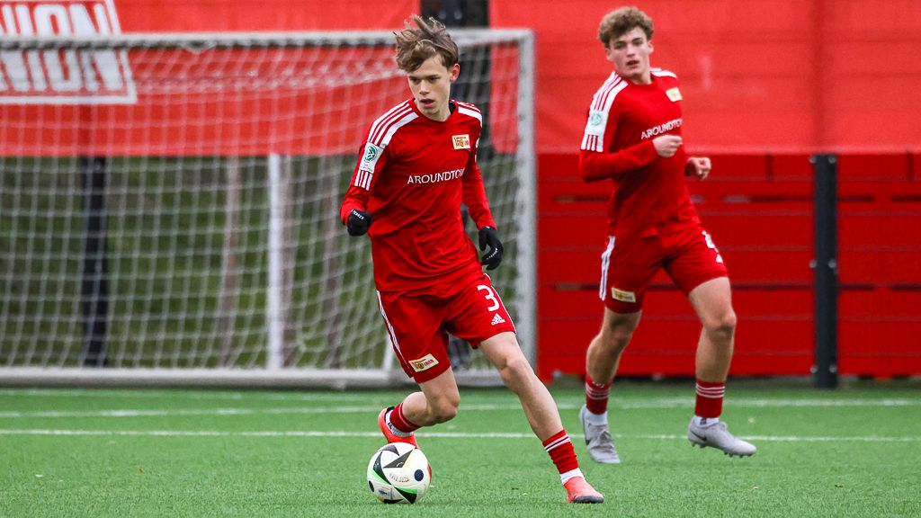 Zwei junge Fußballspieler in roten Trikots trainieren auf einem Kunstrasenplatz. Einer dribbelt den Ball.