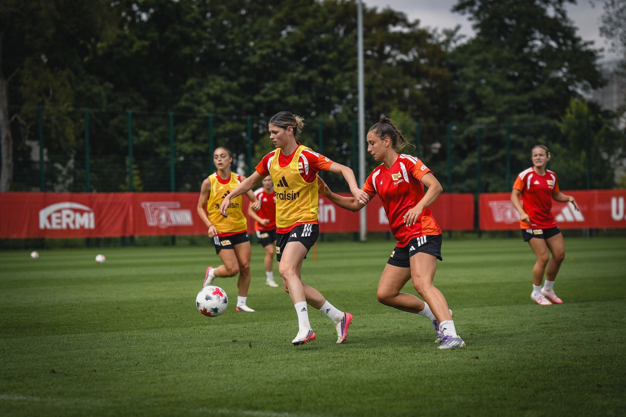 Zwei Fußballspielerinnen dribbeln den Ball auf einem Trainingsplatz, umgeben von weiteren Spielerinnen im Hintergrund.