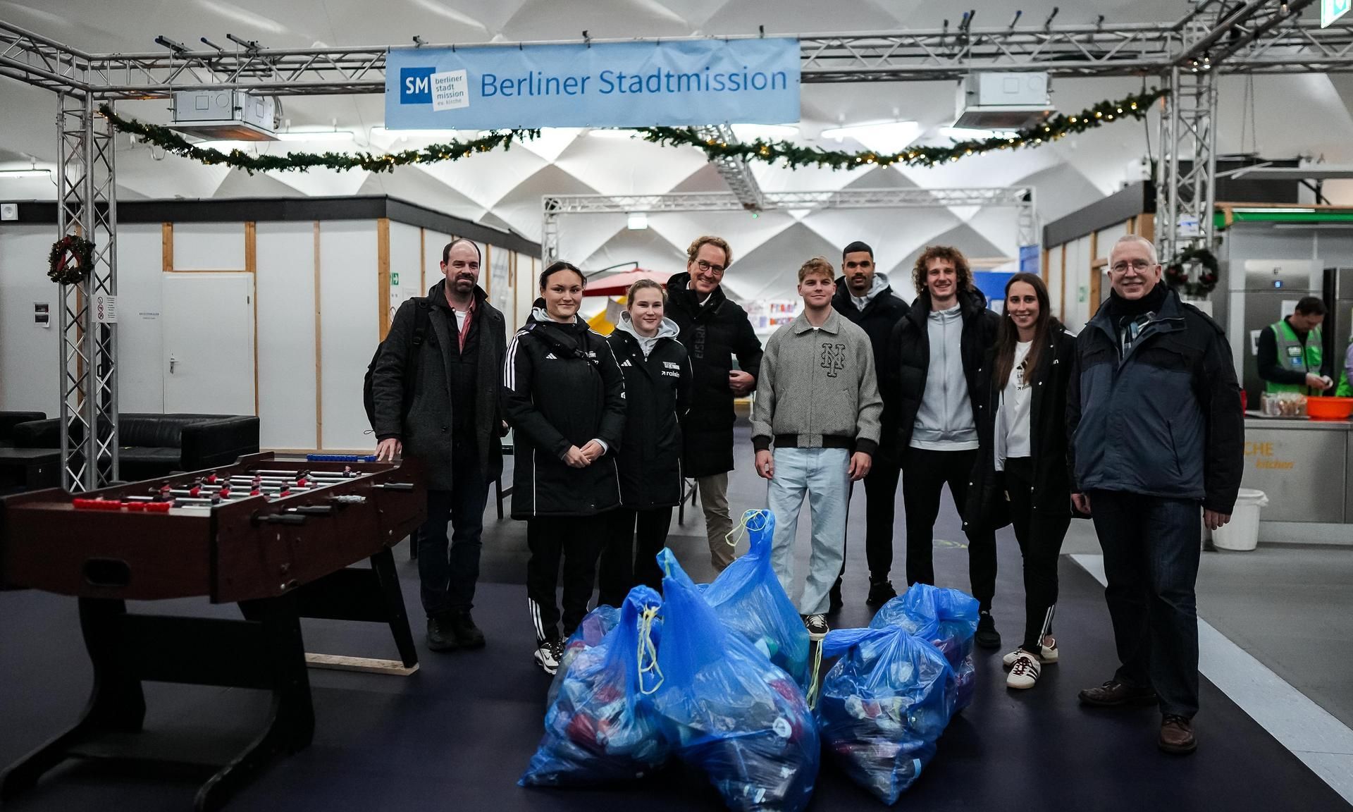 Gruppe von Menschen steht lächelnd vor einem Banner der Berliner Stadtmission, mit blauen Säcken voller gesammelter Gegenstände.