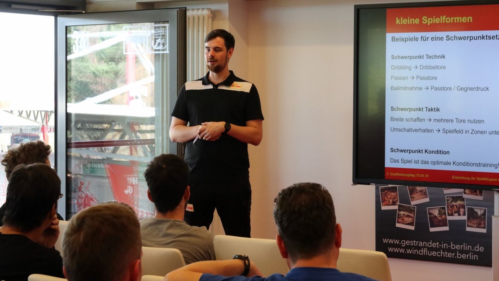 Ein Mann steht vor einer Gruppe von Personen und präsentiert Informationen zu verschiedenen Spielformen im Sport.