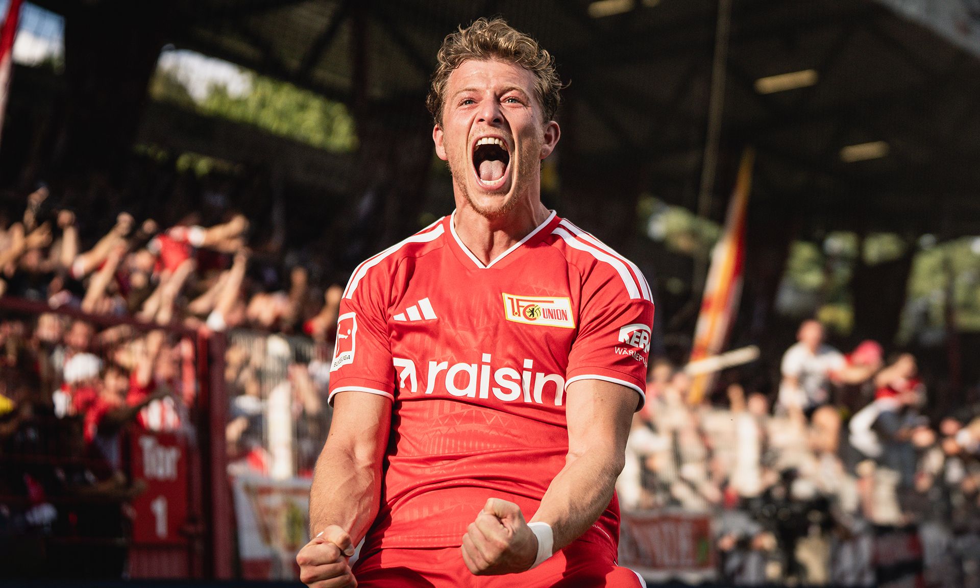 Ein jubelnder Fußballspieler in rotem Trikot feiert einen Torerfolg vor jubelnden Fans im Stadion.
