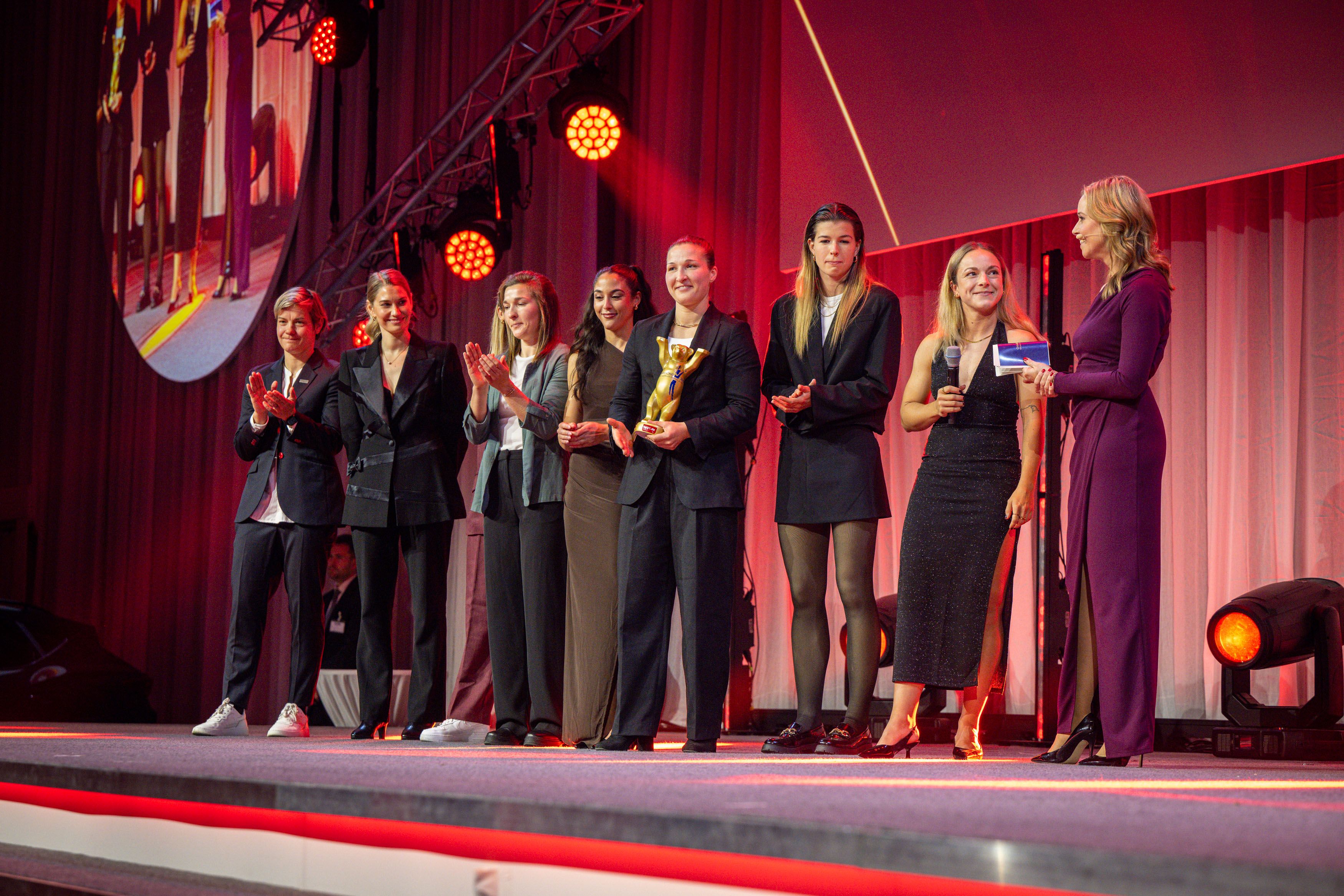 Sieben Frauen stehen auf einer Bühne, applaudieren und halten Trophäen, während eine Moderatorin spricht. Beleuchtung in warmen Tönen.