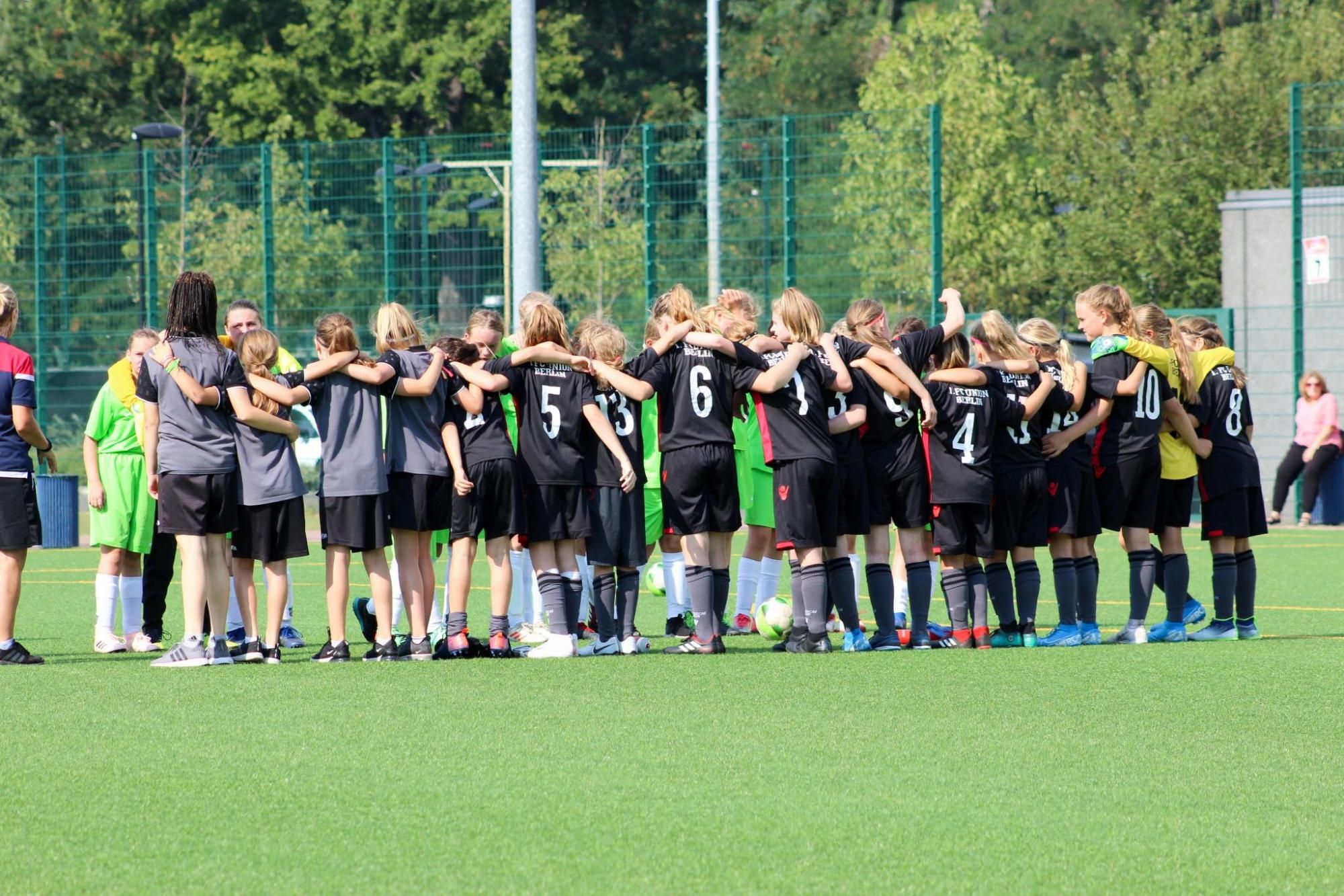 Gruppe junger Fußballspielerinnen in schwarzen Trikots steht umarmt auf dem Spielfeld, einige Zuschauer im Hintergrund.