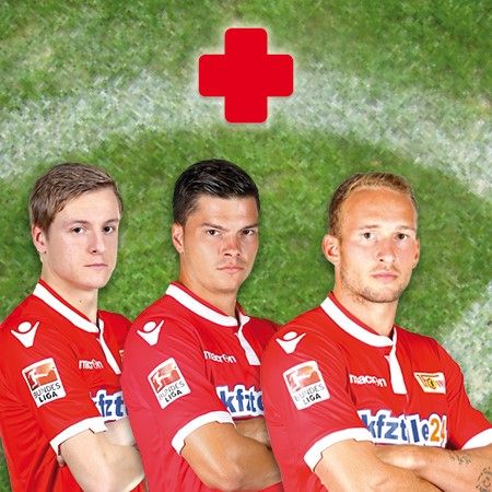 Drei Fußballspieler in roten Trikots stehen nebeneinander, vor einem grünen Hintergrund mit einem roten Kreuz darüber.