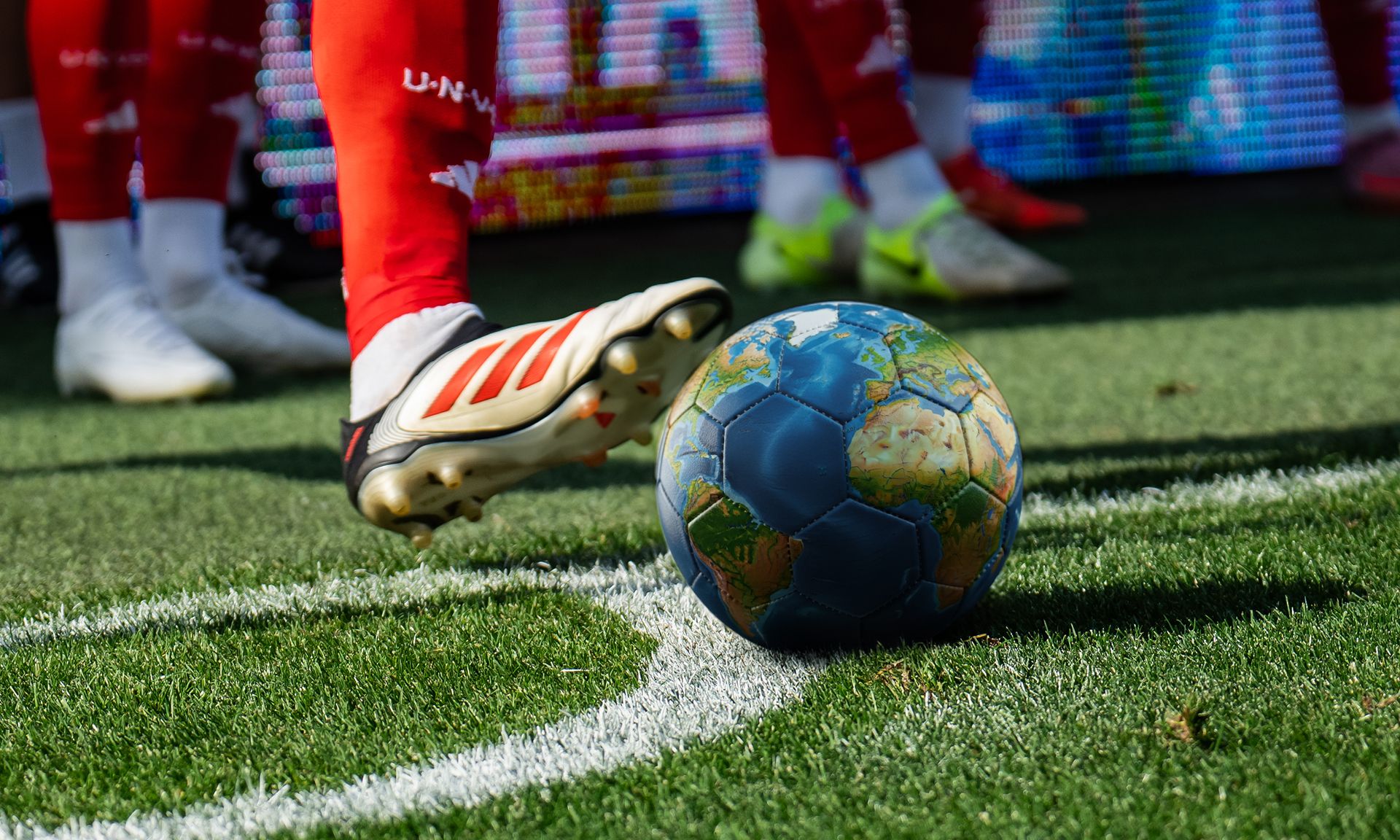 Ein roter Fußballschuh berührt einen erdigen Fußball auf einem Spielfeld mit Kunstrasen.