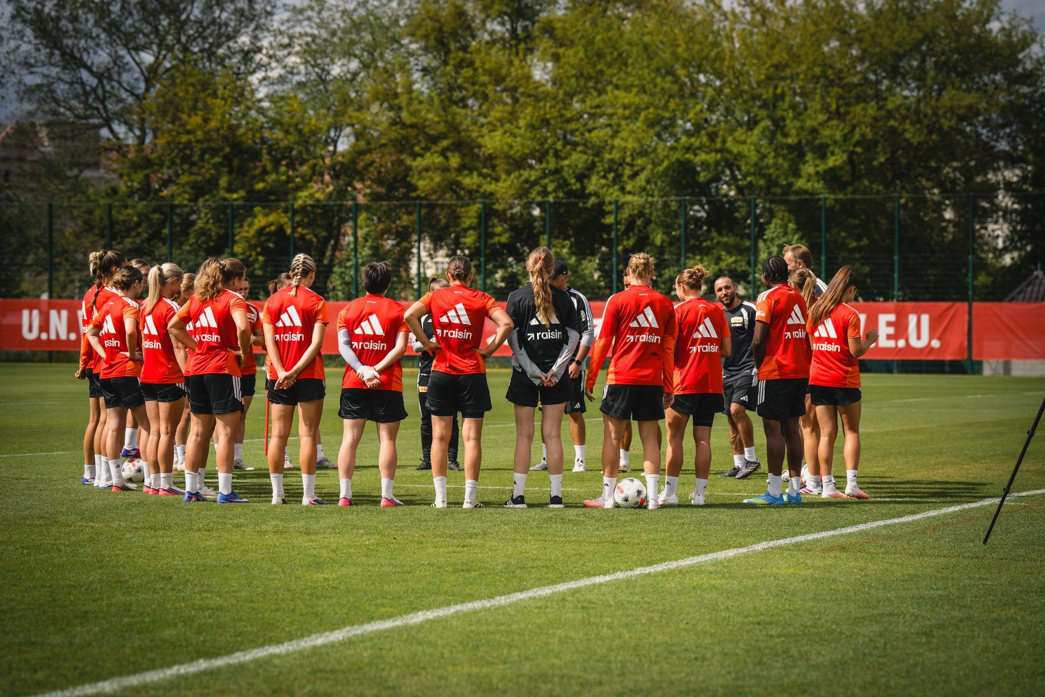 Eine Gruppe von Fußballspielerinnen im roten Trainingsdress steht im Kreis auf einem Rasenplatz und hört einem Trainer zu.
