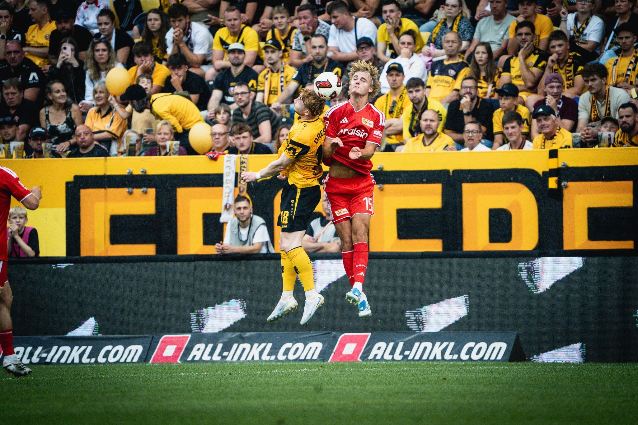 Zwei Fußballspieler springen in der Luft, um den Ball zu kämpfen. Im Hintergrund ist eine jubelnde Menge zu sehen.