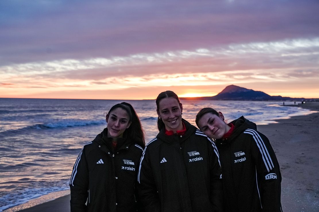Drei Personen in Sportjacken stehen am Strand, während die Sonne hinter ihnen untergeht und den Himmel in sanften Farben erleuchtet.