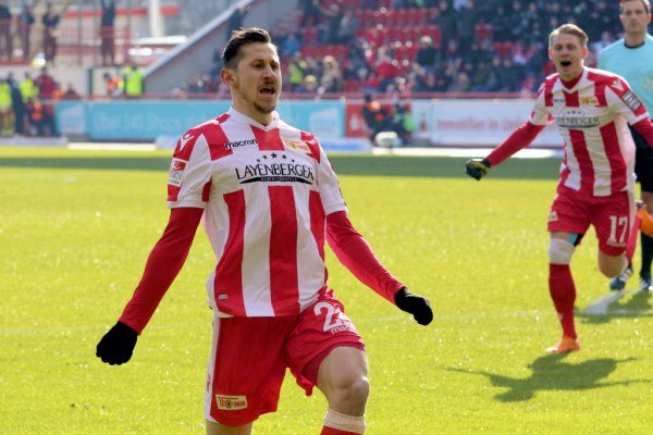 A soccer player in a red and white jersey celebrates his goal while a teammate cheers in the background.