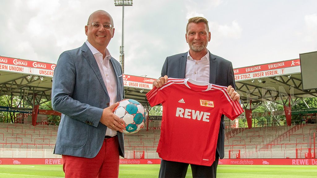 Two men are standing in the stadium, one holding a soccer ball and a jersey that says "REWE."